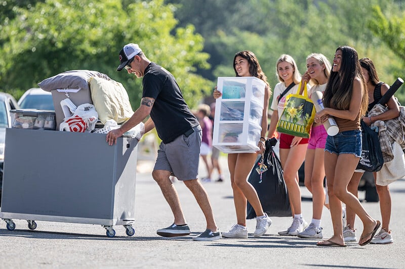 A family member pushes a cart of student belongings across a parking lot on Move-In Day at UM