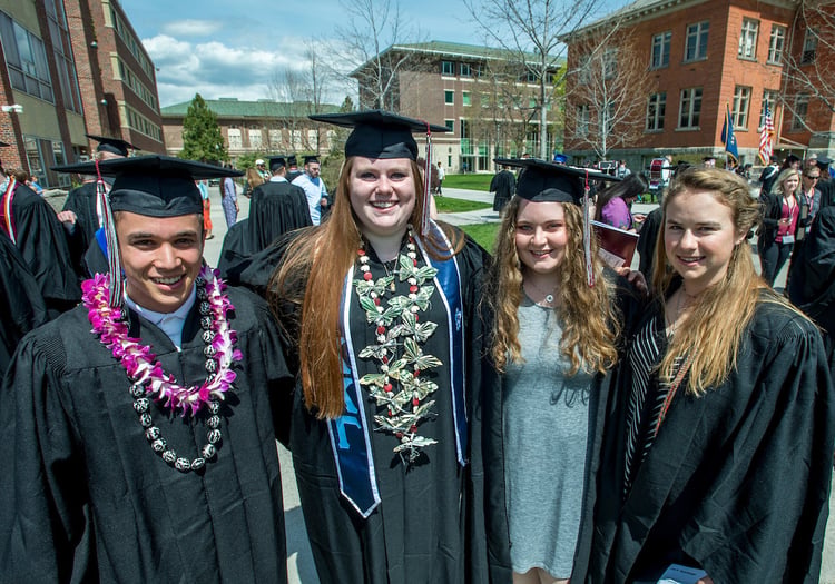 Four graduates in caps and gowns stand outside and smile toward the camera