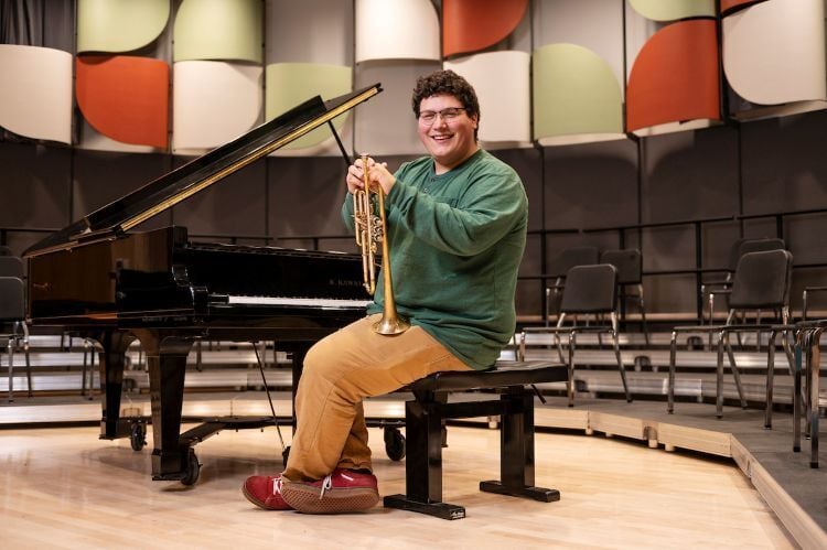 A music major sitting in front of a piano with his trumpet