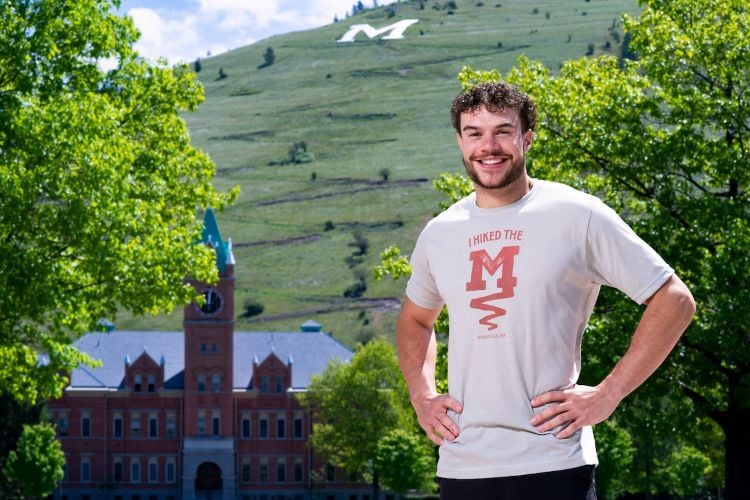 UM Football player stands below Mount Sentinel wearing a I Hiked the M Trail