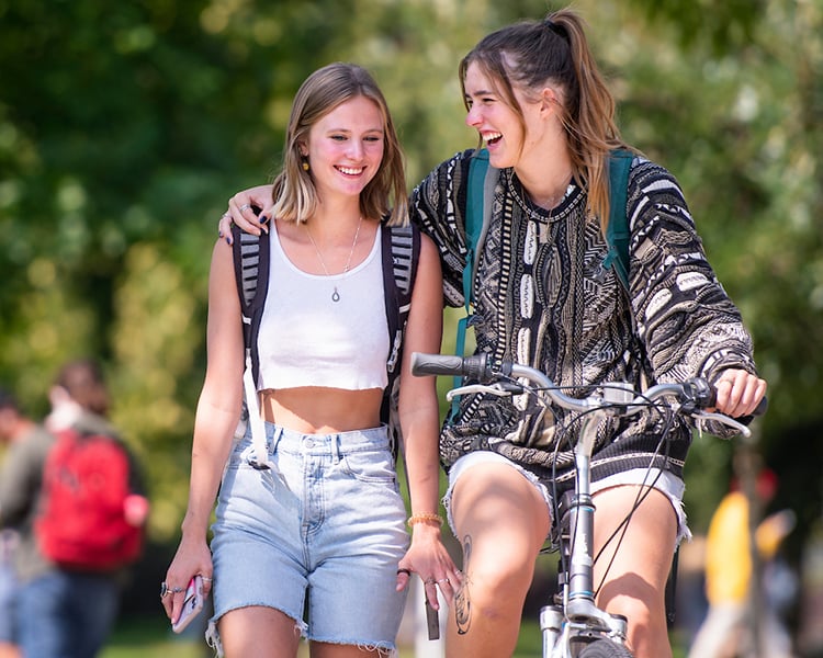 a girl on a bike and a girl next to her. girl on bike has arm around her friend.