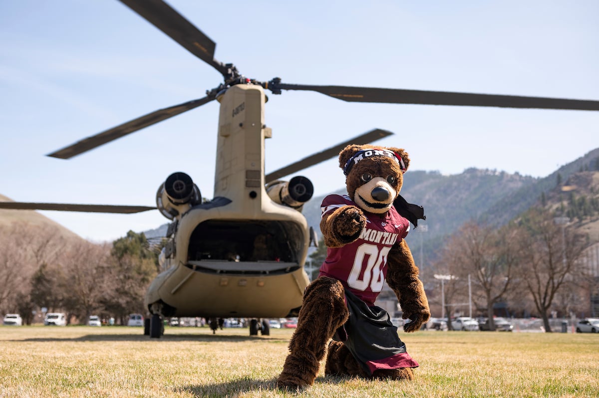 Monte the mascot gives a confident pointing finger with a military helicopter in the background.