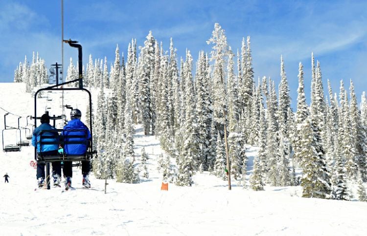 Skiers in blue jackets sit side-by-side on a chairlift at Lost Trail Powder Mountain on a blue bird ski day.
